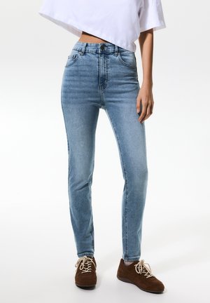 Femme portant un jean skinny taille haute bleu clair, un t-shirt court blanc et des baskets à lacets marron, se tenant devant un fond blanc uni.