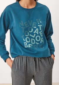 Felpa color turchese con lettere stampate testurizzate che formano la frase "NON È MAI TROPPO TARDI PER FARE MEGLIO", abbinata a pantaloni jogger grigi con cordoncino.