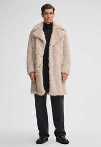 Uomo in piedi che indossa un cappotto in pelliccia sintetica beige sopra una maglietta nera con zip, pantaloni neri e scarpe nere, davanti a uno sfondo chiaro uniforme.