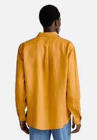 Camicia a maniche lunghe in lino giallo senape brillante; presenta un colletto standard, polsini con bottone e un orlo curvo, con una vestibilità rilassata.