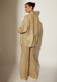 Lichtbeige loungewear set van een gestructureerde, glanzende stof. Het oversized shirt heeft lange mouwen en zijsplitten; bijpassende wijde broekspijpen maken de look compleet.