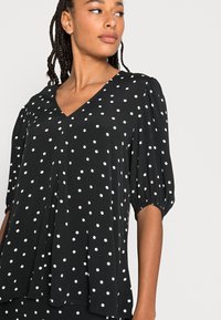 Camisa preta com bolinhas brancas, decote em V e mangas curtas bufantes. Corte solto com pregas na frente. Tecido suave e leve.