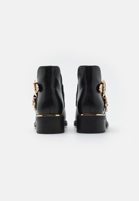 Botas de tobillo de cuero negro con un tacón bajo, que presentan un acento decorativo en tono dorado en el lateral. Textura suave, diseño elegante.