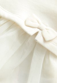 Weißes Kleidungsstück mit einem weichen Stoffoberteil mit Schleifenakzent und einem mehrlagigen Tüllrock, der eine zarte transparente Textur zeigt.