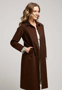 Manteau long marron en tissu à texture lisse ; doté d'un col, d'une fermeture à boutons et de poignets à carreaux.