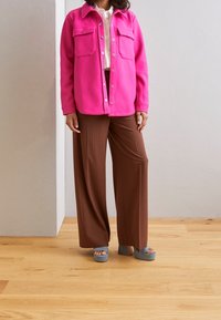 Růžová fleece bunda se dvěma předními kapsami, kombinovaná s hnědými pruhovanými širokými kalhotami a světle modrými platformovými sandály.