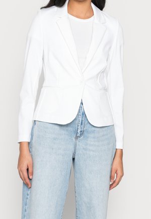 Femme portant un blazer blanc ajusté par-dessus un top blanc et un jean taille haute bleu clair sur un fond uni.