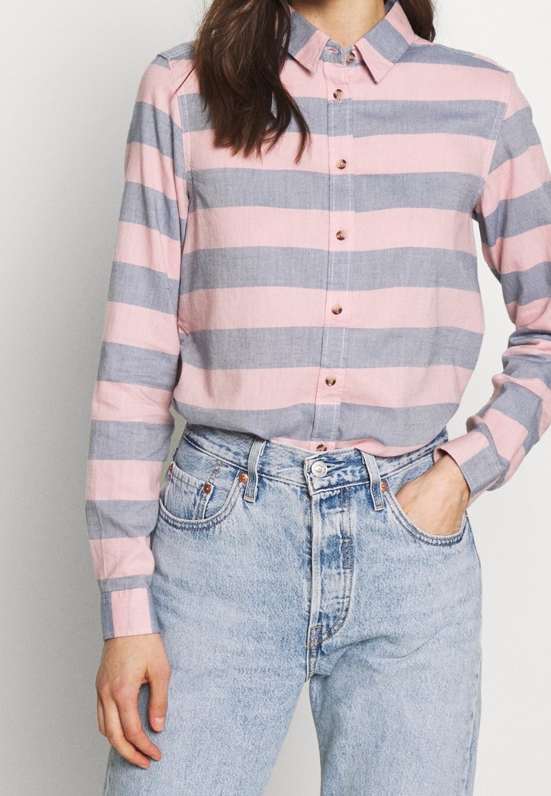 Femme portant une chemise boutonnée à rayures horizontales roses et grises, rentrée dans un jean taille haute bleu clair avec une main dans la poche.