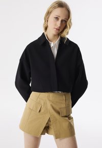 Schwarze Jacke mit Kragen und offenem Frontteil, kombiniert mit beigen Shorts, die zwei Fronttaschen und einen ausgefransten Saum haben.