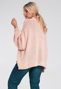 Jasnoróżowy sweter dziany o luźnym kroju, z prążkowanymi mankietami i okrągłym dekoltem, w połączeniu z ciemnymi dżinsami, widoczny z boku.