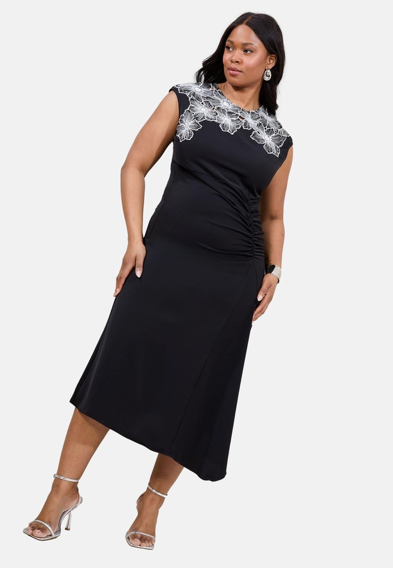 Rochie neagră cu un corset ajustat, mâneci scurte și broderie florală la decolteu. Prezintă o parte plisată și o fustă fluidă.