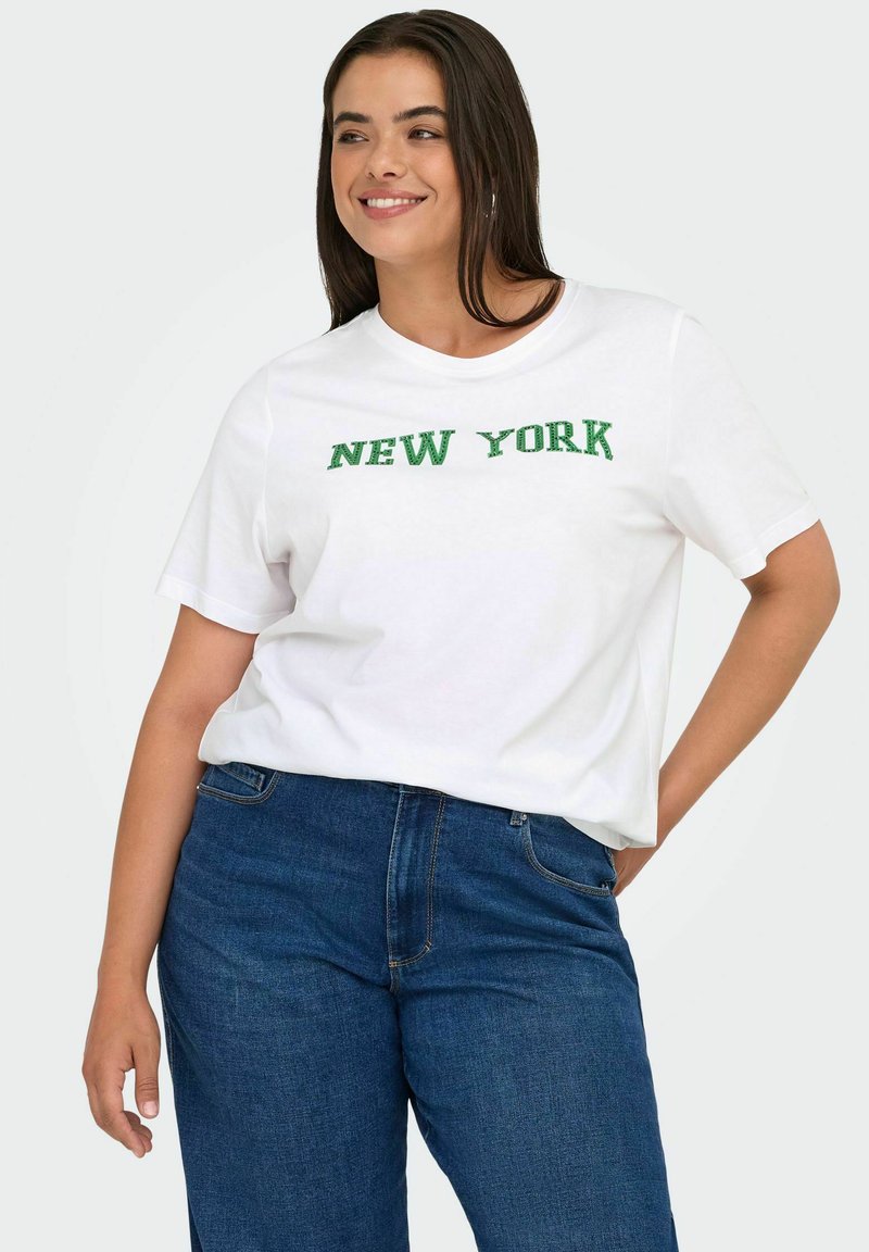 Moteris šypsosi, vilkinti baltą marškinėlį su žodžiu "NEW YORK" žaliais rašmenimis ir mėlyniais džinsais, stovinti su viena ranka kišenėje.