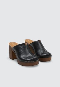 Zuecos negros de cuero sin cordones con gruesos tacones plataforma de madera y costuras visibles, mostrados sobre un fondo gris claro liso.