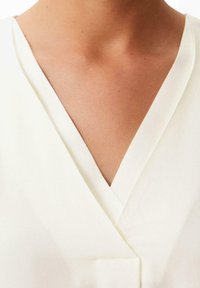 Blouse couleur crème avec encolure en V, présentant un tissu lisse, une légère brillance et des lignes épurées ; aucun motif ni ornement visible.