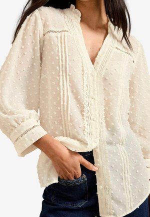 Kvinde iført en hvid tekstureret bluse med puffede ærmer og blonde-detaljer, hånd hvilende i mørkeblå flettede jeans med lommer.