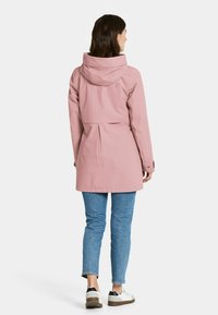 Vaaleanpunainen vedenpitävä takki, jossa on säädettävä huppu, kiristettävä vyötärö ja takana laskostus. Yhdistettyinä sinisiin farkkuihin ja valkoisiin lenkkareihin.