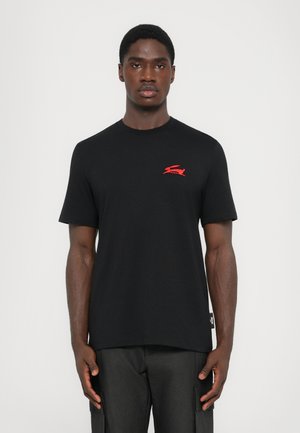 Black cotton t-shirt with short sleeves, featuring a red logo on the left chest and a side tag detail. Smooth texture, relaxed fit.