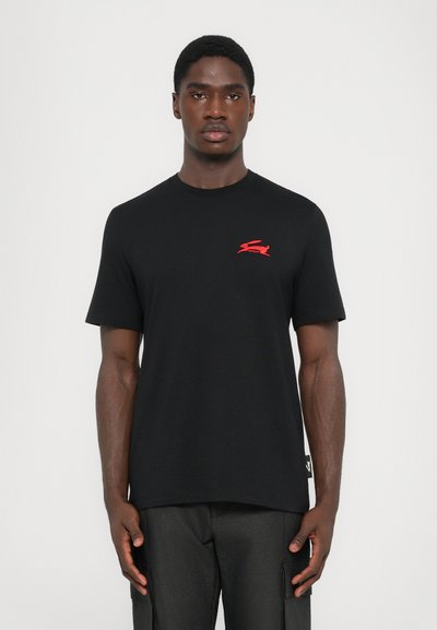 T-shirt en coton noir à manches courtes, avec un logo rouge sur la poitrine gauche et un détail d'étiquette sur le côté. Texture lisse, coupe décontractée.