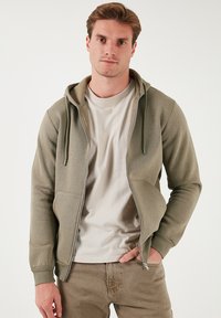 Sudadera con cremallera en verde oliva, con capucha ajustable y bolsillos frontales, y puños de canalé, llevada sobre una camisa beige clara, combinada con pantalones marrón claro.