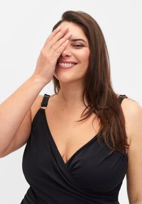 Maillot de bain noir avec un design enveloppant, des bretelles ajustables, une texture douce et une finition lisse, porté par une personne qui se couvre le visage d'une main.