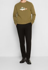 Muž v olivově zelené mikině s potiskem ryby, černých kalhotách a černých kožených botách, stojící s rukama v kapsách.