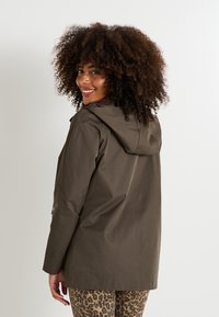 Chaqueta impermeable verde oliva con capucha, cintura ajustada y mangas largas, combinada con leggins de estampado de leopardo. Textura suave y diseño minimalista.