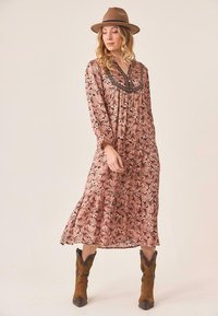 Mujer vistiendo un vestido midi floral rosa con mangas largas, un sombrero marrón y botas vaqueras de ante marrón, de pie sobre un fondo neutro.