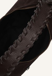 Sangle en cuir brun foncé avec des coutures décoratives sur un sac en suède brun foncé partiellement ouvert avec des dents de fermeture éclair en laiton visibles.