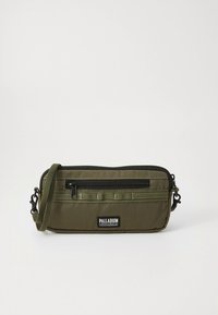 Bolsa transversal retangular verde-oliva com bolso frontal com fecho de correr, acabamento preto, alça ajustável e etiqueta da marca Palladium na frente.