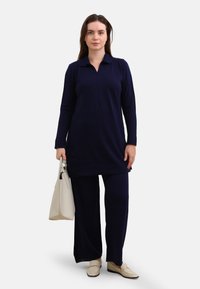 Navy blue tunic with a collar and long sleeves, paired with matching pants. Model holds a beige tote bag. Cream-colored slip-on shoes.