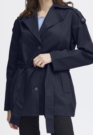 Laivansinisellä vyöllä varustettu trench-coat, jossa on leveä kauluksen, nappikaitale, sivutaskut ja olkatoppaukset, valmistettu sileästä kankaasta.