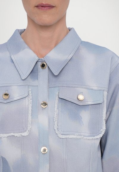 Lys blå denimjakke med sky-lignende mønstre, to frontlommer, frynset kanter og gullfarvede knapper med sorte centre.