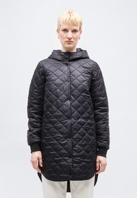 Άτομο με κοντά ξανθά μαλλιά, φοράει μαύρο καπιτονέ μπουφάν με κουκούλα και κουμπιά ασφαλείας, καθώς και ανοιχτόχρωμο παντελόνι, στέκεται μπροστά σε απλό φόντο.