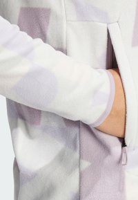 Chaqueta de forro polar en blanco suave con patrones geométricos en lavanda. Cuenta con un bolsillo con cremallera y detalles en puños acanalados. Textura acogedora.