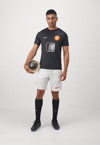 Svart Nike fotballskjorte med prikkemønster, oransje merke, hvit løve-logo. Kombinert med hvite shorts, svarte sokker og svarte fotballsko.
