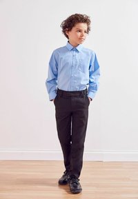 Chemise bleu clair à manches longues avec fermeture à boutons et petite poche poitrine, associée à un pantalon noir et des chaussures noires, debout sur un sol en bois.