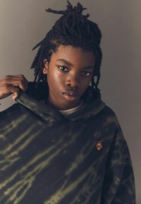 Schwarz-grüner Tie-Dye-Hoodie mit einem orangenen Totenkopf-Logo auf der Brust. Weicher Stoff, oversize Schnitt und eine Kapuze mit Kordelzug.