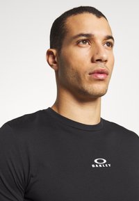 Sort T-shirt lavet af bomuld med rund halsudskæring. Et hvidt "OAKLEY" logo er synligt på brystet. Enkel, ensfarvet design uden mønstre.