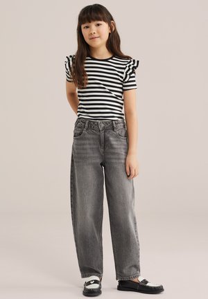 Jong meisje met lang bruin haar, gekleed in een zwart-wit gestreept shirt met ruches aan de mouwen, grijze wijde jeans en zwart-witte loafers.