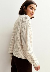 Une jeune femme aux cheveux bruns courts porte un pull en tricot texturé de couleur crème et un pantalon foncé, se retournant sur son épaule devant un mur uni.