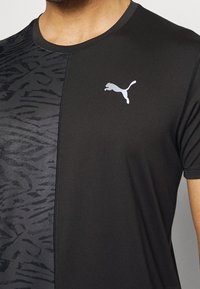 Camiseta atlética negra con un panel texturizado a la izquierda y un panel liso a la derecha. Presenta un pequeño logo blanco de Puma en el pecho.