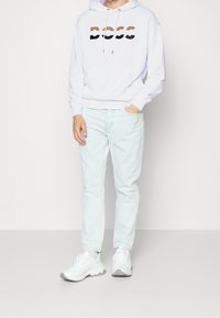 Sudadera blanca con el logotipo "BOSS" en marrón y negro, jeans azul claro y zapatillas blancas con detalles en gris. Diseño de atuendo casual.