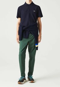 Marinblå pikétröja med en liten logotyp, gröna byxor och en marinblå kabelstickad tröja knuten i midjan. Svarta sneakers med färgade accenter.