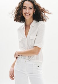 Camisa blanca de manga corta con botones y dos bolsillos frontales, hecha de una tela ligera. Combinada con vaqueros blancos que presentan detalles en dorado.