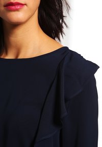 Blusa blu navy con dettagli a volant sulle spalle, realizzata in tessuto morbido, con scollatura rotonda e maniche corte. Vista ravvicinata del colletto.