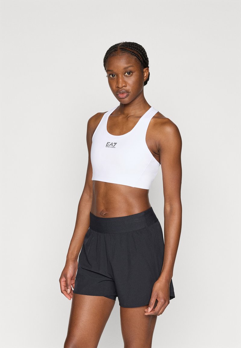 Λευκό αθλητικό σουτιέν με στρογγυλή λαιμόκοψη και λογότυπο στο κέντρο, συνδυασμένο με μαύρο σορτς με ελαστική μέση και χαλαρή εφαρμογή.