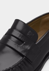 Mocassin penny en cuir noir avec une lanière décorative et un petit logo carré embossé près de l'ouverture de la chaussure sur un fond blanc.