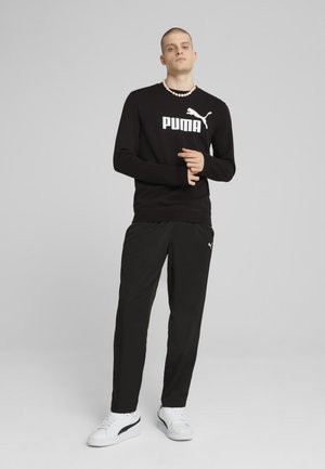 Čierna mikina s bielym logom "PUMA", kombinovaná so voľnými čiernymi nohavicami. Model má na sebe biele tenisky s modrými akcentmi a stojí na neutrálnom pozadí.