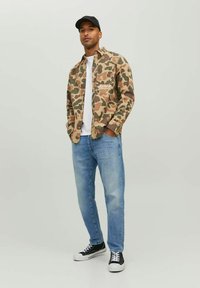 Chaqueta de camuflaje con cierre de botones y logo blanco en el pecho, camiseta blanca, jeans de mezclilla azul claro y zapatillas negras con suelas blancas.