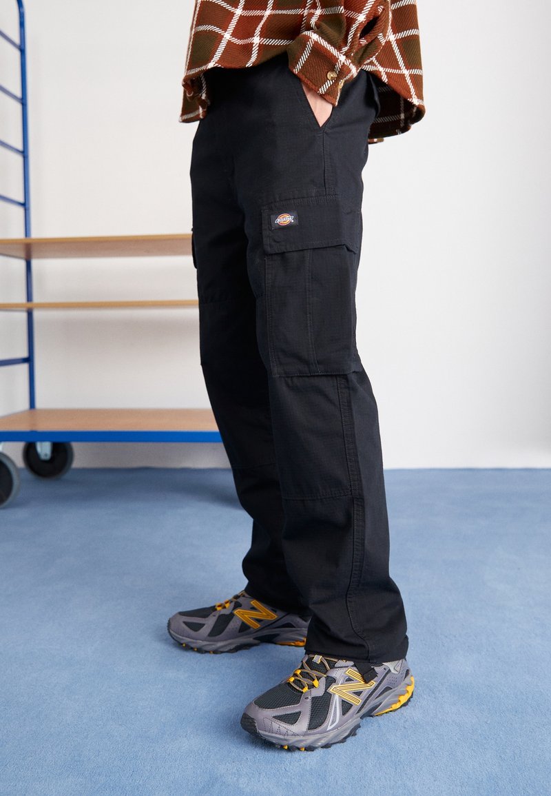 Czarne spodnie cargo z bocznymi kieszeniami, zestawione z szarymi butami sportowymi z żółtymi akcentami i fakturowaną powierzchnią.